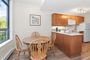 A kitchen with wooden cabinets, a white refrigerator, a microwave, and a dining table with chairs. - Alpenglow Lodge Bear Wonder One-Bedroom by MVA (Whistler)
