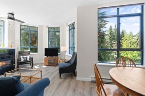 A modern living room with a fireplace, large windows, a TV, and a dining area. - Alpenglow Lodge Bear Wonder One-Bedroom by MVA (Whistler)