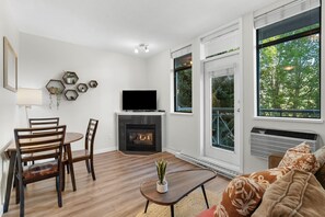 Ein Wohnzimmer mit Kamin, einem Esstisch mit Stühlen, einer Couch und großen Fenstern mit Blick auf Bäume.