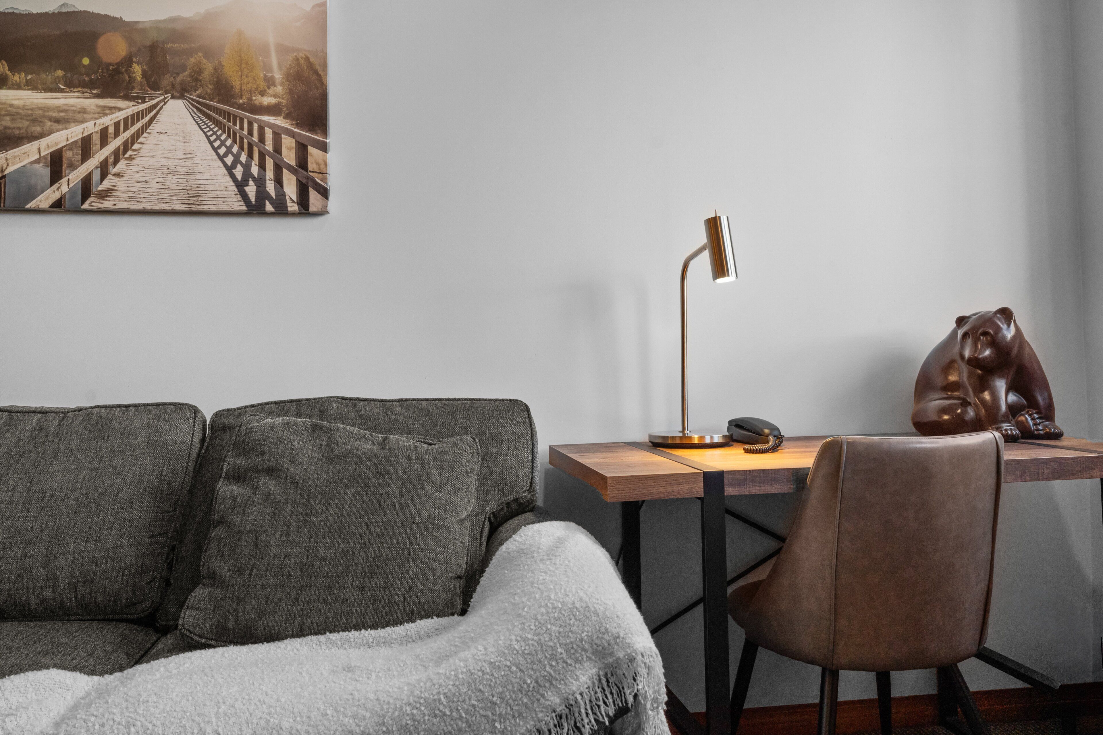 Una stanza con un divano grigio, una scrivania in legno con una lampada, una statua di un orso e un quadro di un ponte.