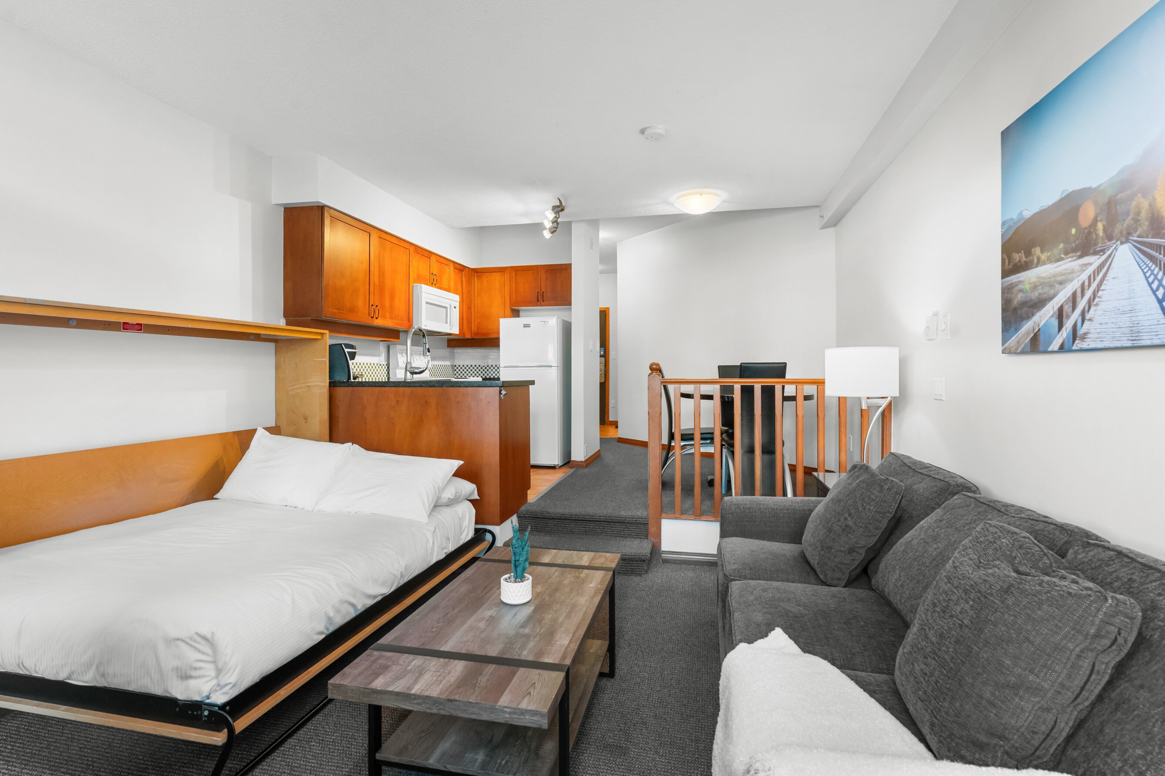Quarto de hotel compacto com cama, sofá, mesinha, cozinha compacta e um quadro na parede.