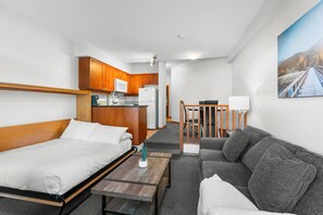 A compact hotel room with a bed, a sofa, a small table, a kitchenette, and a painting on the wall.