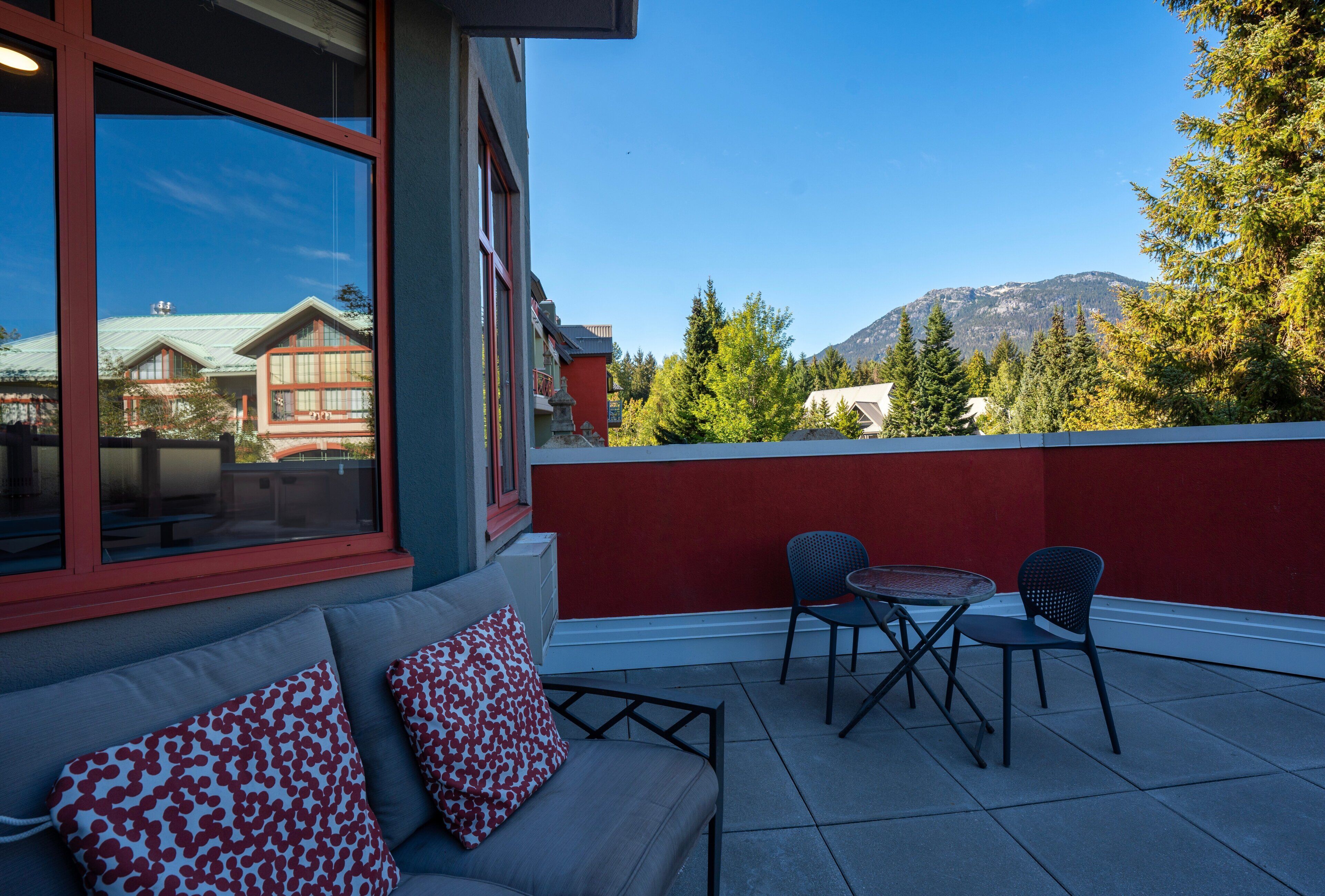 A balcony with a red wall, a gray sofa with patterned cushions, and two chairs.