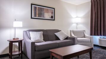 A hotel room with a grey sofa, two patterned armchairs, a wooden coffee table, a side table with a lamp, and a framed artwork on the wall.