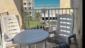 A balcony with a round table and two chairs, overlooking a beach and a building with balconies.