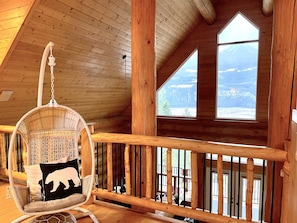 A wooden cabin interior with a high ceiling, a hanging chair with a bear pillow, and large windows offering a view of a mountain landscape. - Spectacular mountain views in beautiful log chalet (Golden)