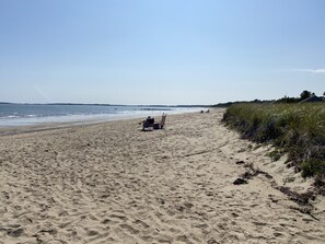 A wide sandy beach with a few beach chairs and umbrellas, a grassy dune area, and a clear view of the ocean.