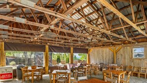 Een eetruimte met houten tafels en stoelen, een hoog plafond met zichtbare balken en grote ramen die uitzicht bieden op het omliggende landschap.