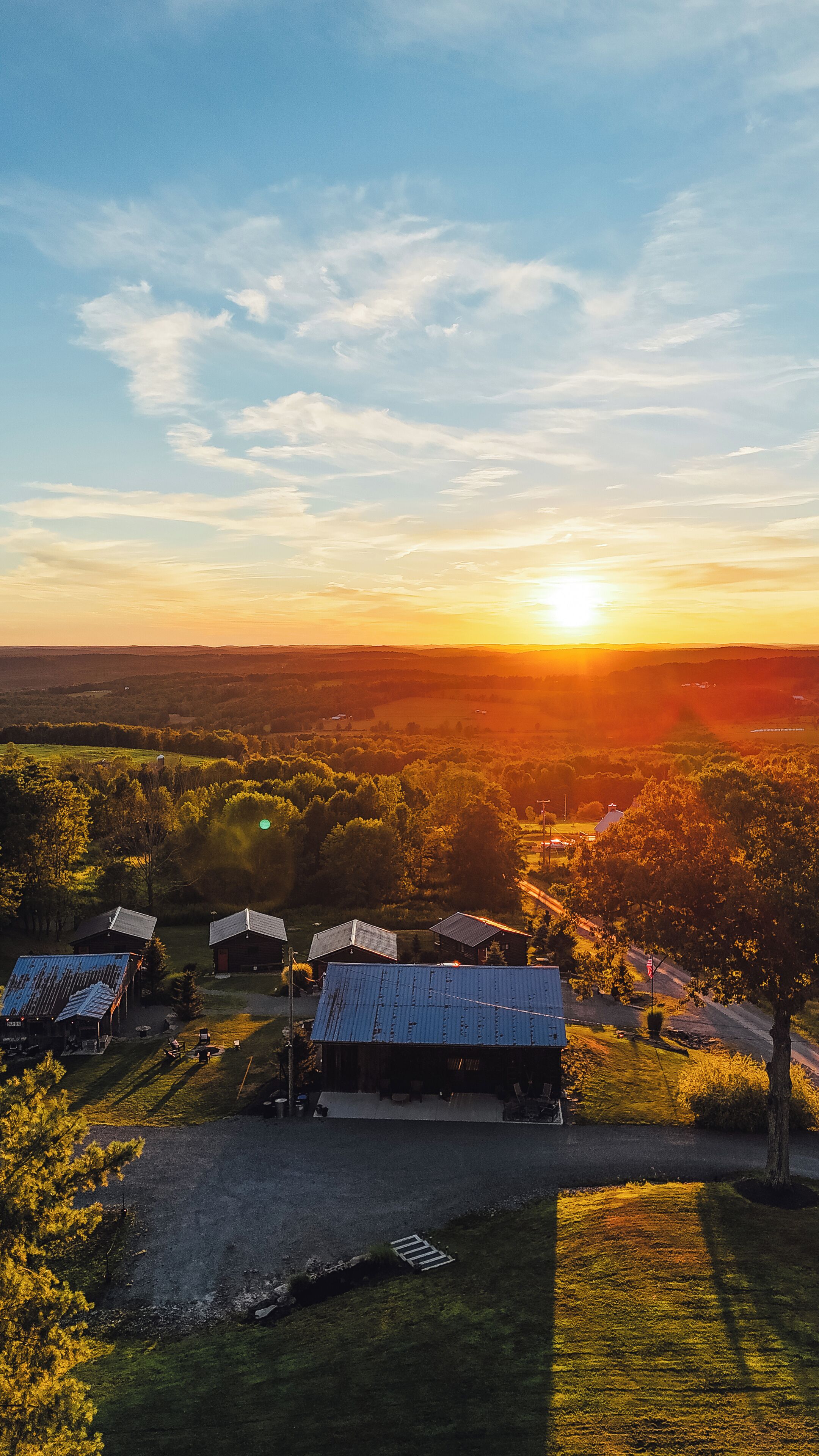 Un paysage rural au coucher du soleil, avec plusieurs bâtiments aux toits métalliques, entourés de verdure et de champs ouverts.