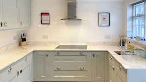 A modern kitchen with light blue cabinets, a stainless steel range hood, and a marble countertop. There are two framed pictures on the wall, a knife block on the counter, and a window with blinds.
