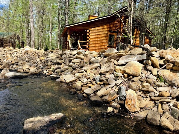 Une cabane en bois, située au bord d’un ruisseau rocheux, entourée d’arbres.