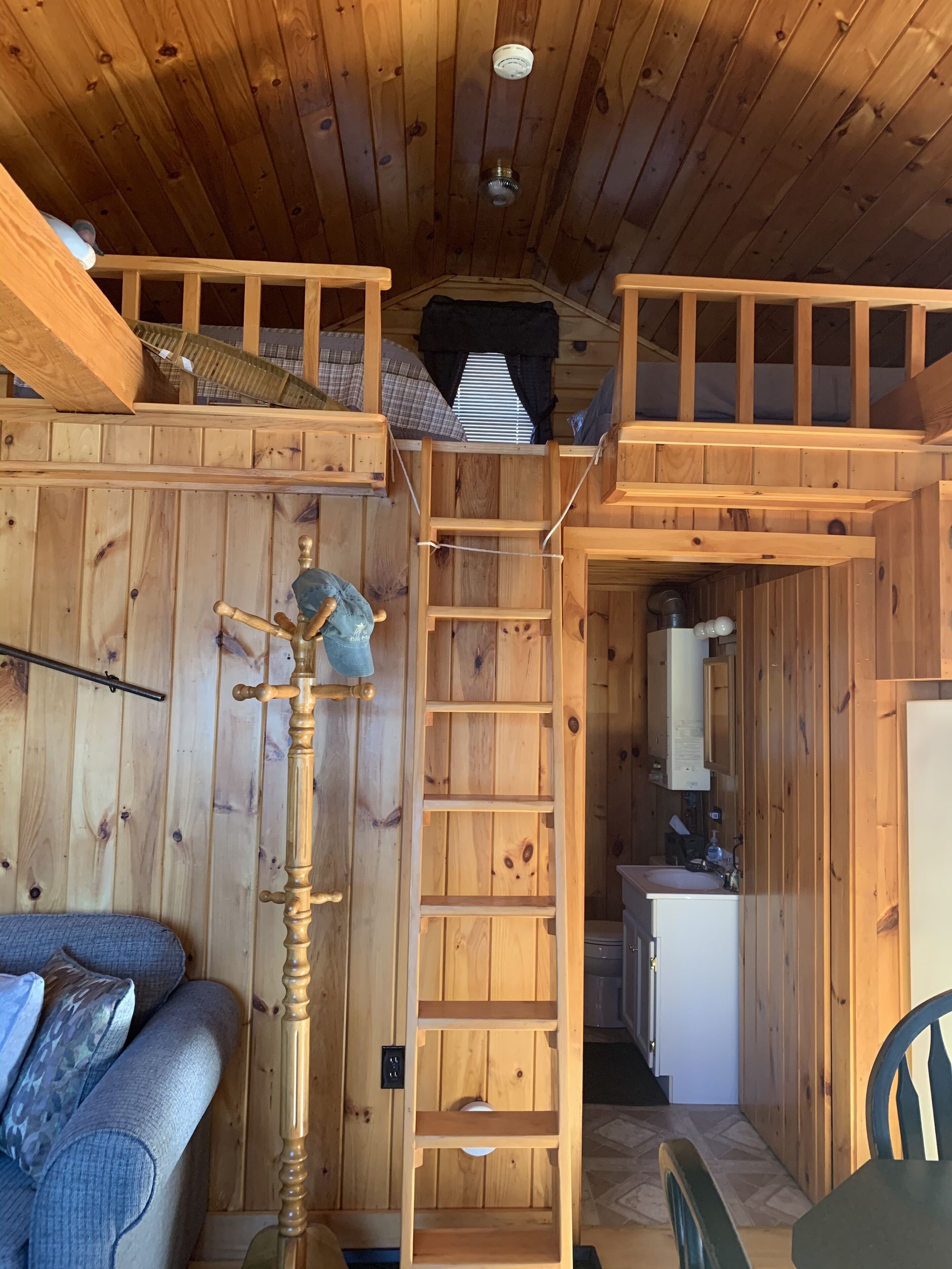 Interior de uma cabana de madeira com cama no mezanino, escada de madeira, pequena cozinha e área de estar com sofá.