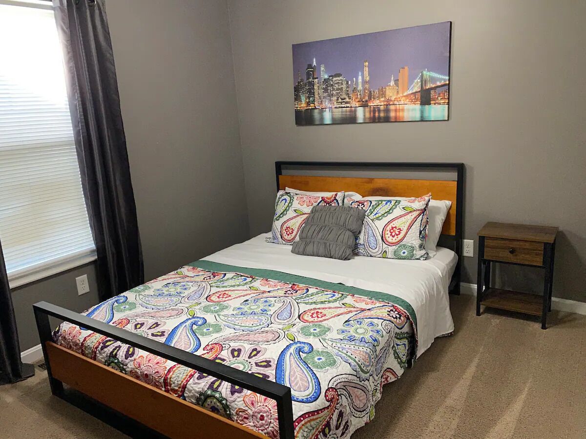 A neatly arranged bedroom with a bed, a nightstand, a window with curtains, and a cityscape painting on the wall.