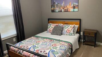 A neatly arranged bedroom with a bed, a nightstand, a window with curtains, and a cityscape painting on the wall.