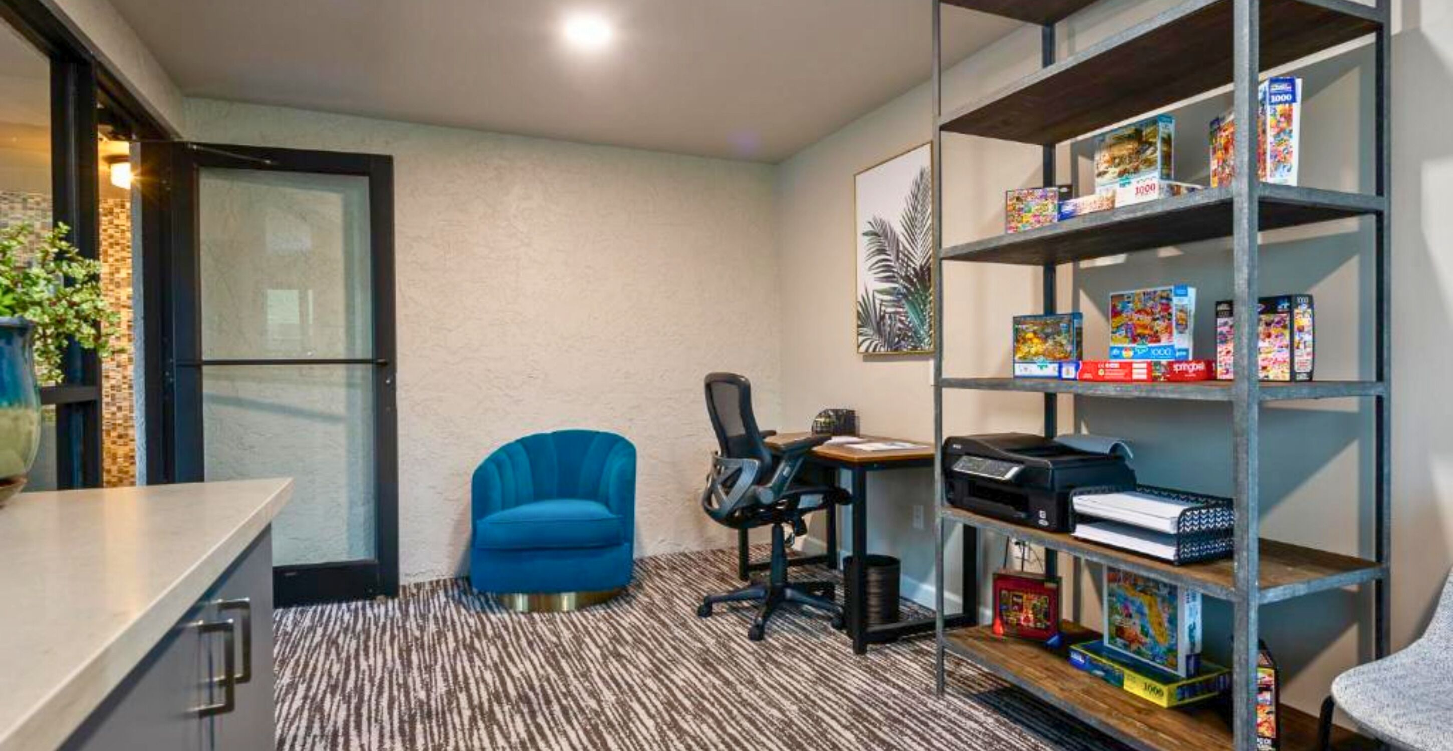 A compact office space with a blue chair, a desk with a printer, and a bookshelf filled with books and games.