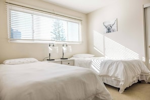 Quarto com duas camas, mesa de cabeceira com luminárias e janela com persianas.