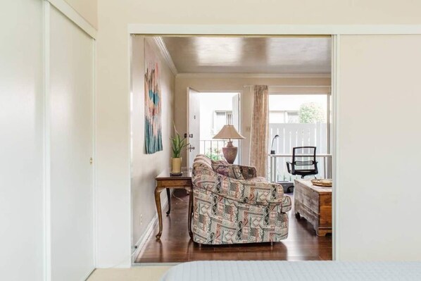 A cozy room with a patterned sofa, a small wooden side table, a lamp, and a view of a balcony.