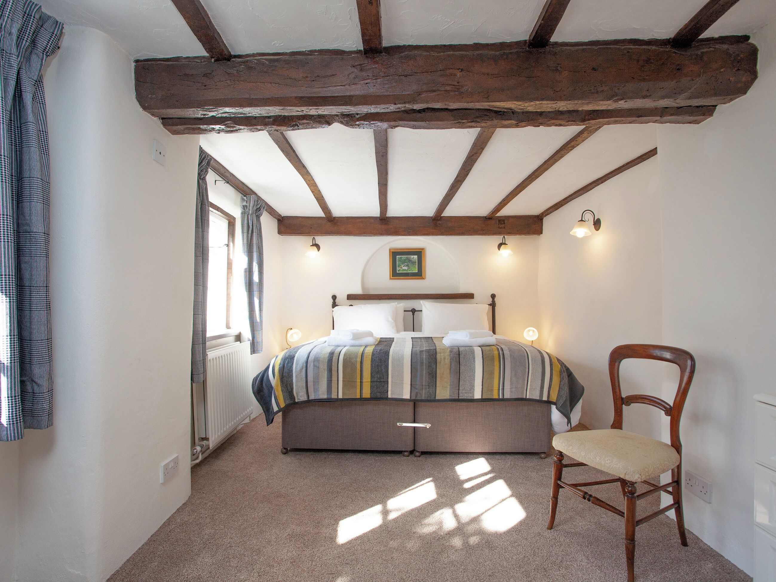 Une chambre à coucher comprenant un lit, une chaise, une fenêtre et un plafond avec des poutres apparentes.
