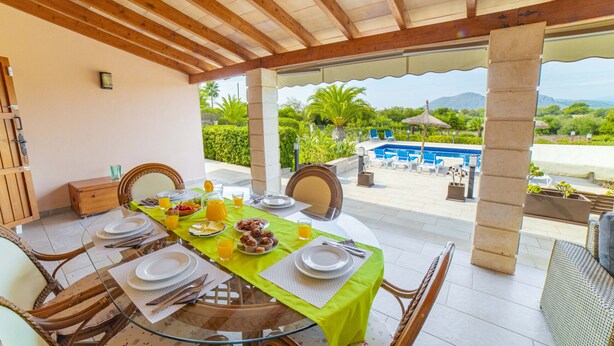 A covered outdoor dining area with a table set for breakfast, overlooking a pool and lush greenery. - Villa Ca na Quarta (Alcúdia)