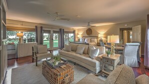 A spacious living room with a large sofa, armchairs, and a coffee table. There are multiple windows providing natural light and views of the outdoors.