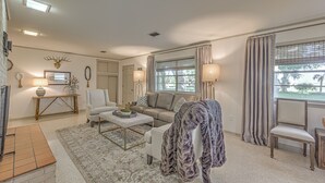 A well-lit living room with a sofa, armchair, and a coffee table. There is a large window with curtains, a floor lamp, and a decorative wall piece.