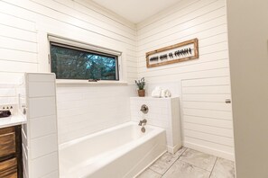 Un bagno con pareti piastrellate di bianco, una finestra, una vasca da bagno, un portascopevoli e una piccola pianta.