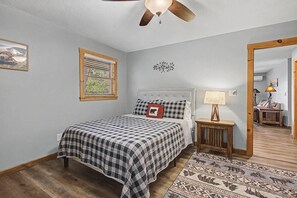 A bedroom with a bed, a bedside table with a lamp, a window with blinds, a ceiling fan, and a rug.