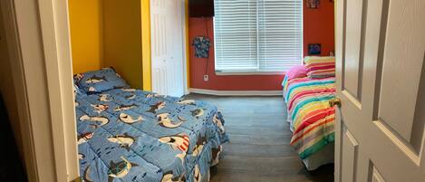 A narrow hallway with two beds, one with a blue shark-themed bedspread and the other with a rainbow-striped bedspread.