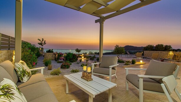Een terras met buitenmeubelen, een tafel en uitzicht op zee bij zonsondergang.