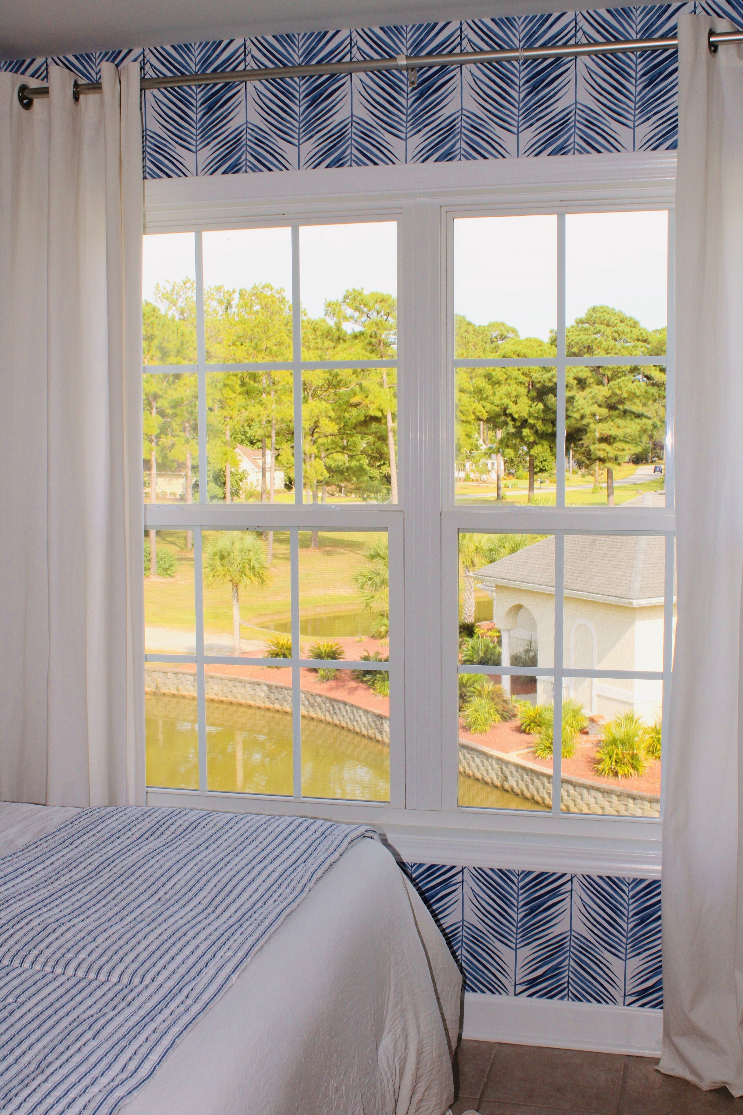A bedroom with a large window, a bed with a striped bedspread, and a view of a garden and pond outside.