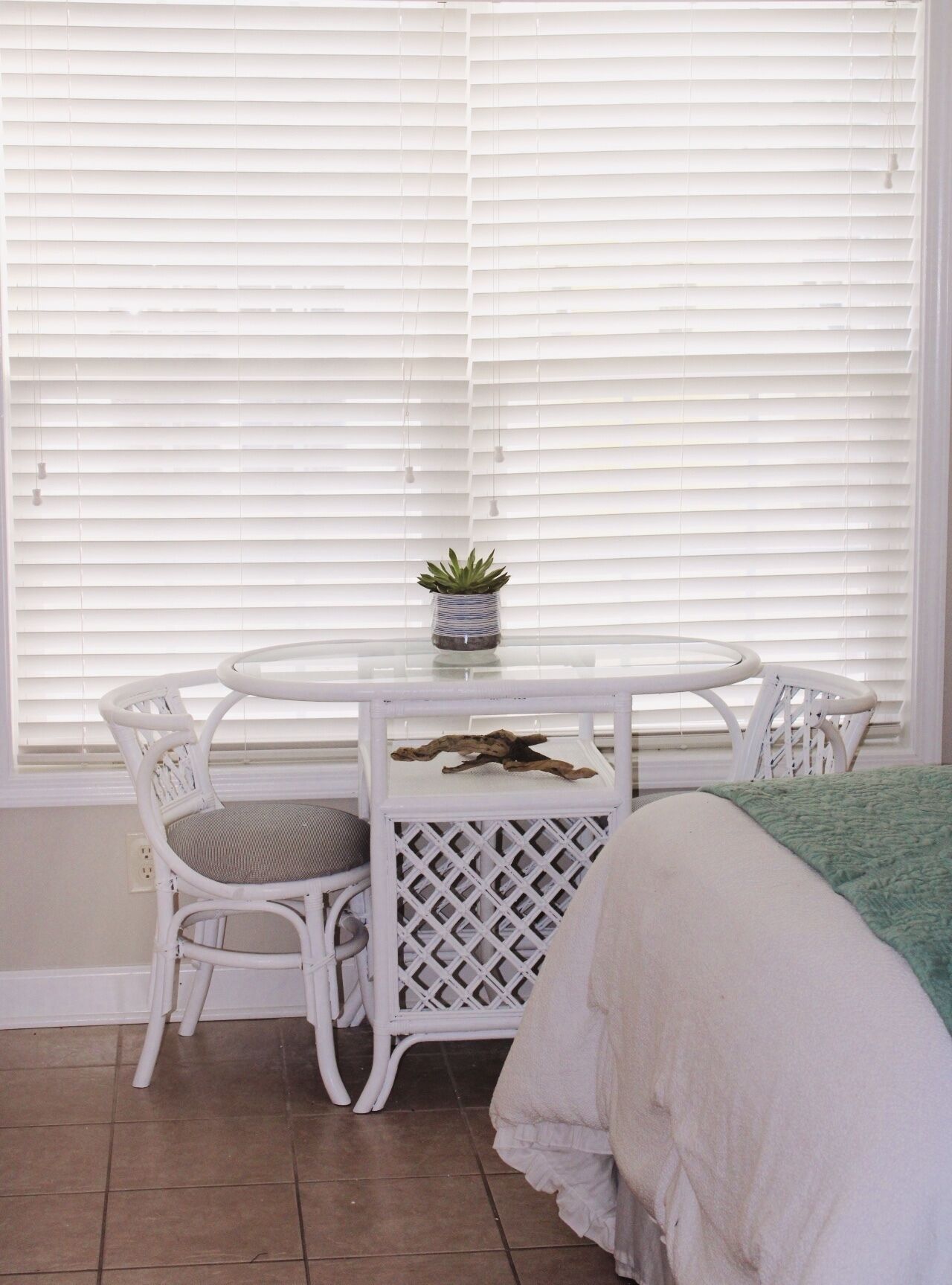 A small, round, white table with a single chair, a potted plant, and a piece of driftwood. The room has white blinds and a green blanket on the bed.