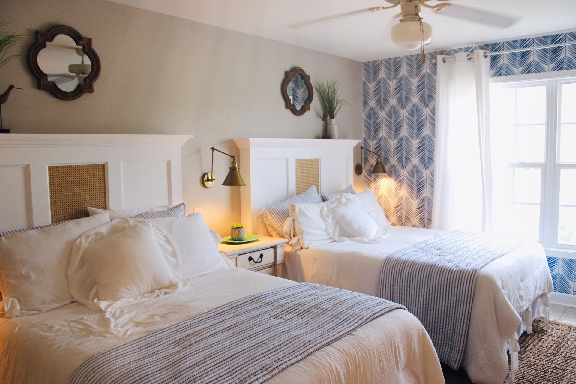 A hotel room with two beds, a ceiling fan, a mirror, a wall clock, and a window with curtains.