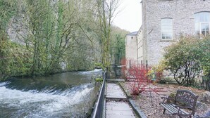 Un edificio de piedra con un río al frente, un sendero y una banca.