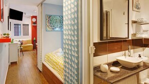 Un baño moderno con un espejo grande, un lavamanos y un inodoro. En el fondo, se ve una cama con una colcha de rayas amarillas y blancas.