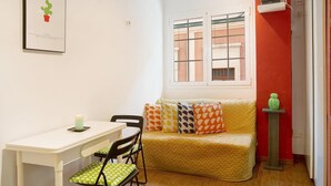 Una habitación pequeña y acogedora con una mesa y sillas blancas, un sofá amarillo con cojines estampados, una ventana con rejas y un cactus verde sobre una mesita.
