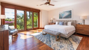 A spacious bedroom with a large bed, a bench, a desk, and a view of greenery through the sliding glass doors.