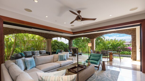 A spacious living area with a large sofa, a coffee table, and a ceiling fan. There is a view of a patio with outdoor furniture and greenery.