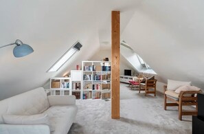 A loft-style room with a sloped ceiling, a white sofa, a wooden bookshelf, and a television.