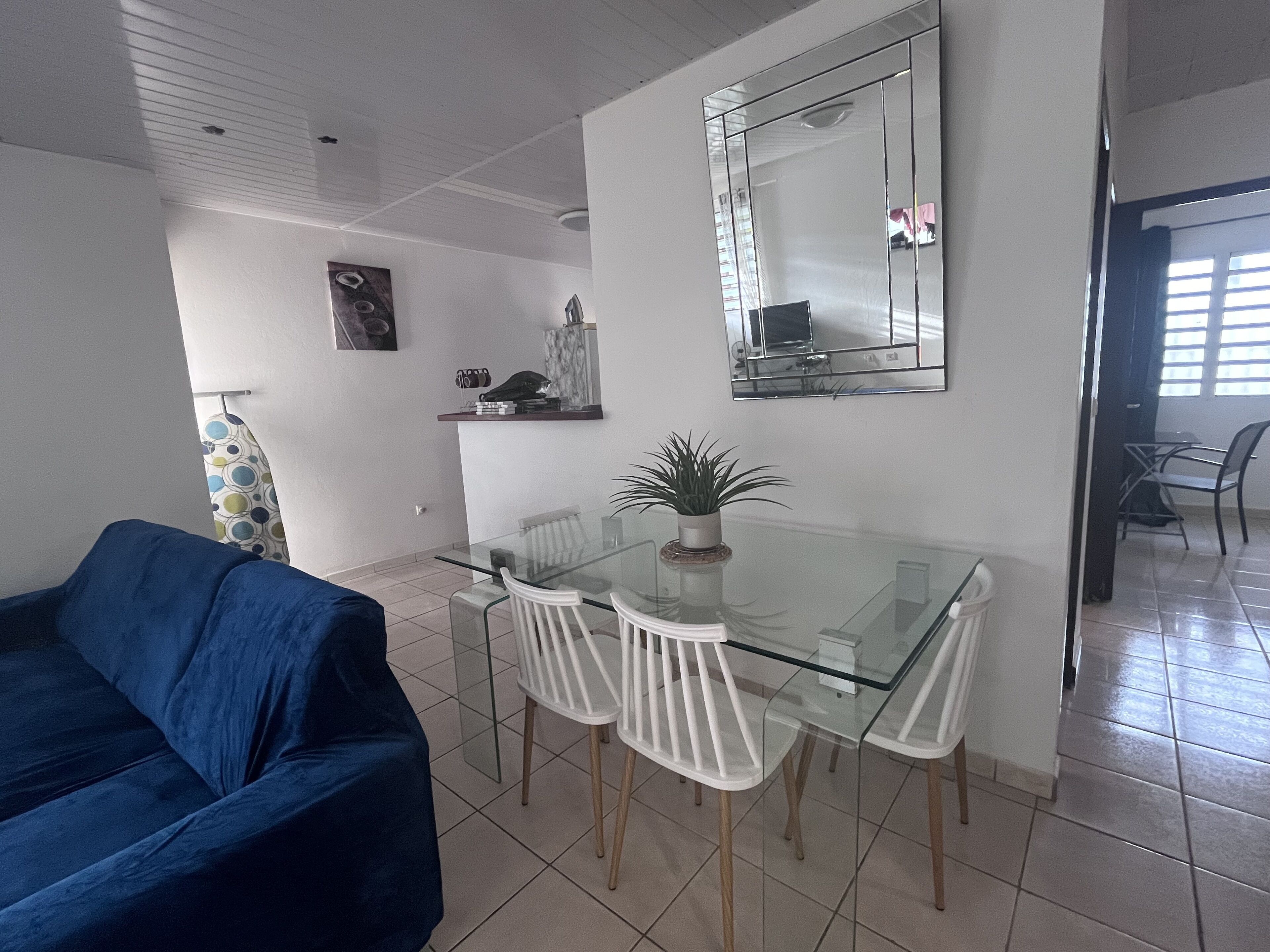 A dining area with a glass table and white chairs, a blue sofa, and a large mirror on the wall.