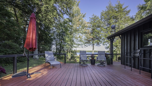 Una terraza de madera con asientos al aire libre, un paraguas rojo y vistas a árboles y un lago.
