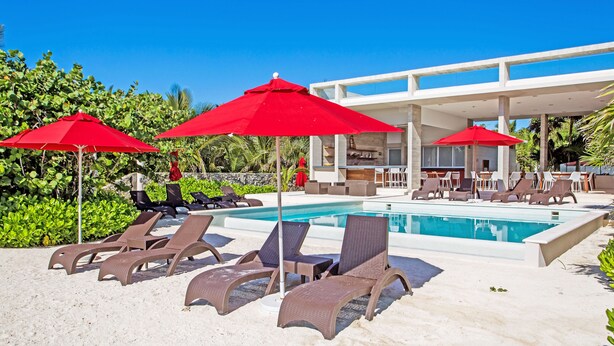 A poolside area with sun loungers and red umbrellas, surrounded by greenery and a modern building structure. - Villa Akumal, A Peaceful Space for 6 with Private Jacuzzi *  Steps to the Beach! (Akumal)