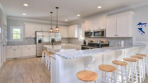 Una cocina moderna con armarios blancos, electrodomésticos de acero inoxidable, una isla de granito con taburetes altos e iluminación colgante.