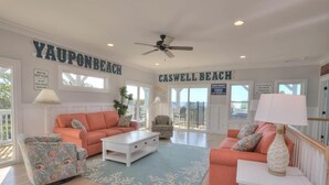 Un salón amplio con dos sofás, una mesa de centro y un sillón estampado. La habitación tiene un ventilador de techo, vistas a la playa y un letrero que dice "Yaupon Beach".