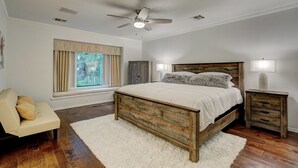 Une chambre à coucher avec un lit en bois, un ventilateur de plafond, une fenêtre avec des rideaux, une table de chevet et une chaise.