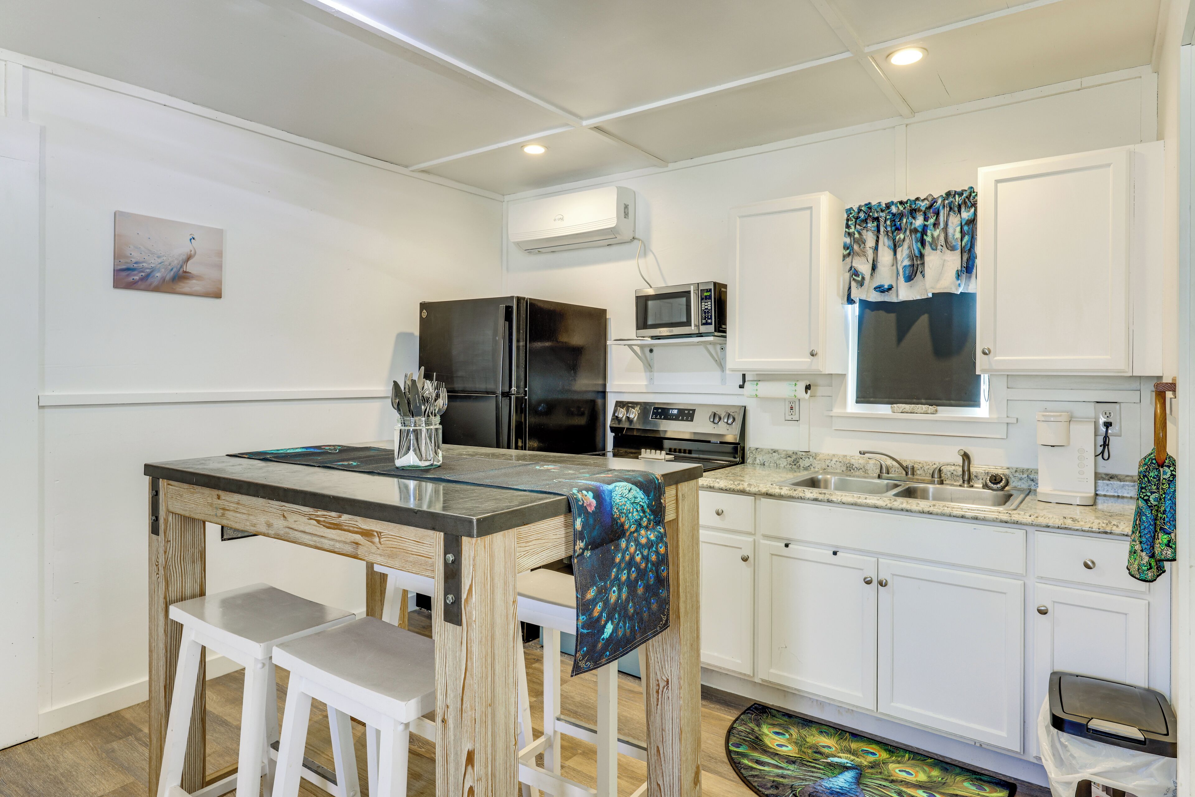 A compact kitchen with white cabinets, a refrigerator, microwave, and a peacock-themed tablecloth.