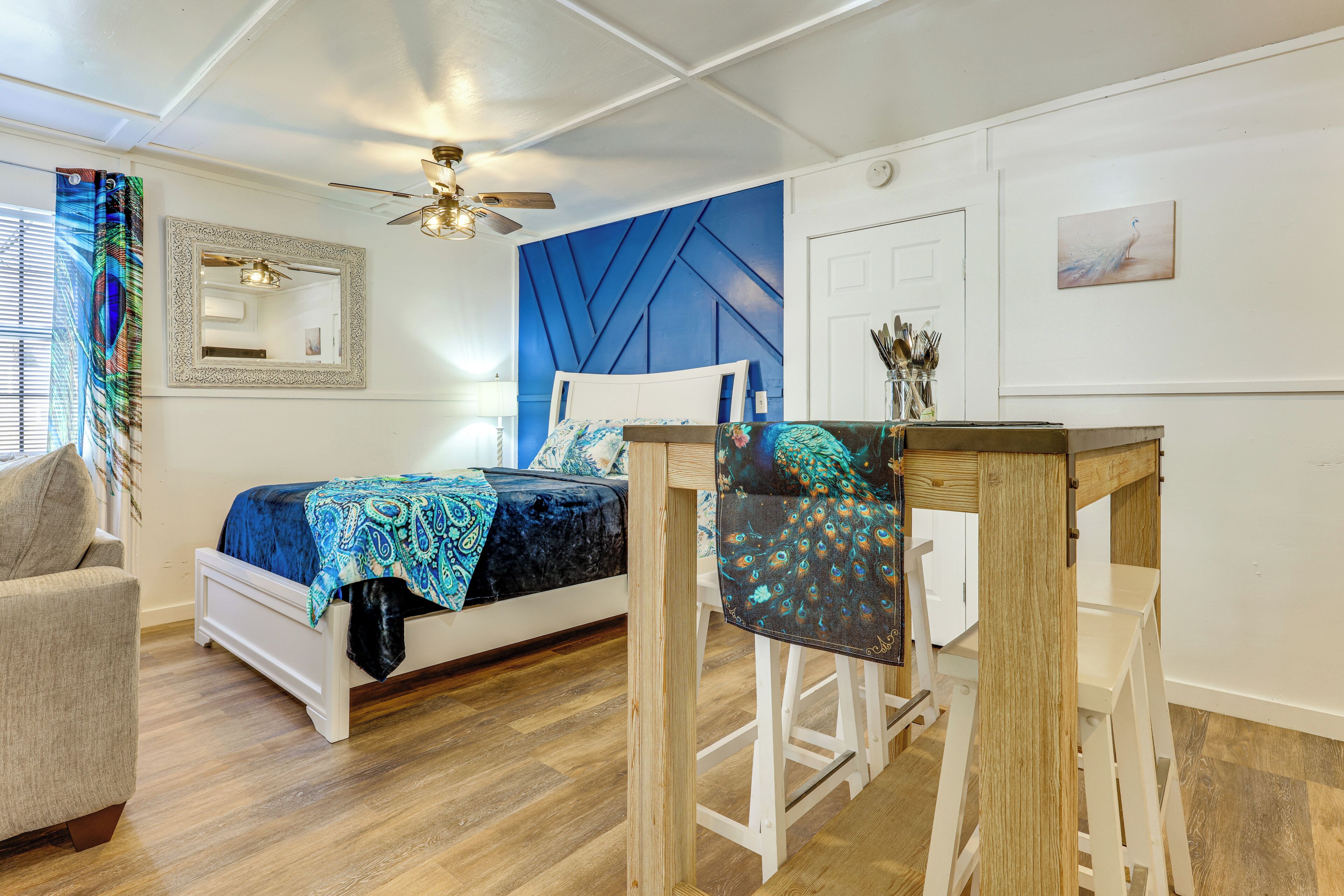 A bedroom with a bed, a chair, a table, a ceiling fan, a mirror, and a door.