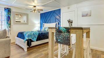 A bedroom with a bed, a chair, a table, a ceiling fan, a mirror, and a door.