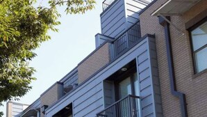 A modern multi-story building with balconies, large windows, and a landscaped area with shrubs.