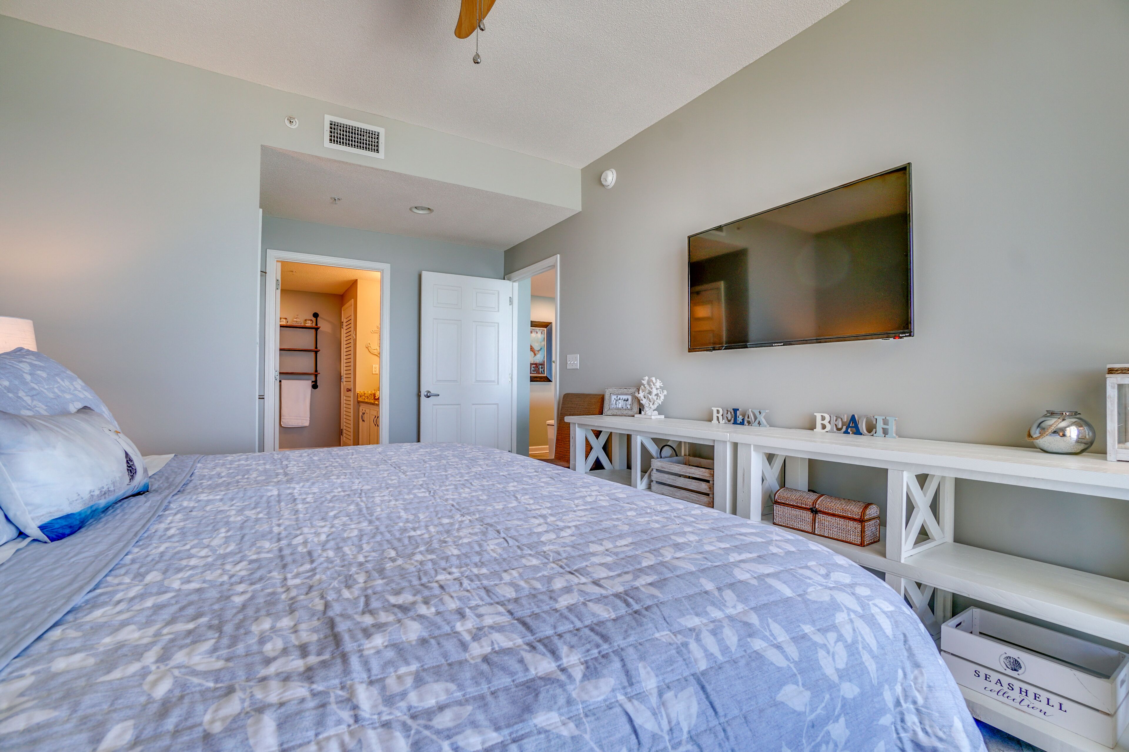 A bedroom with a large bed, a TV mounted on the wall, a desk with a computer, and a bookshelf.