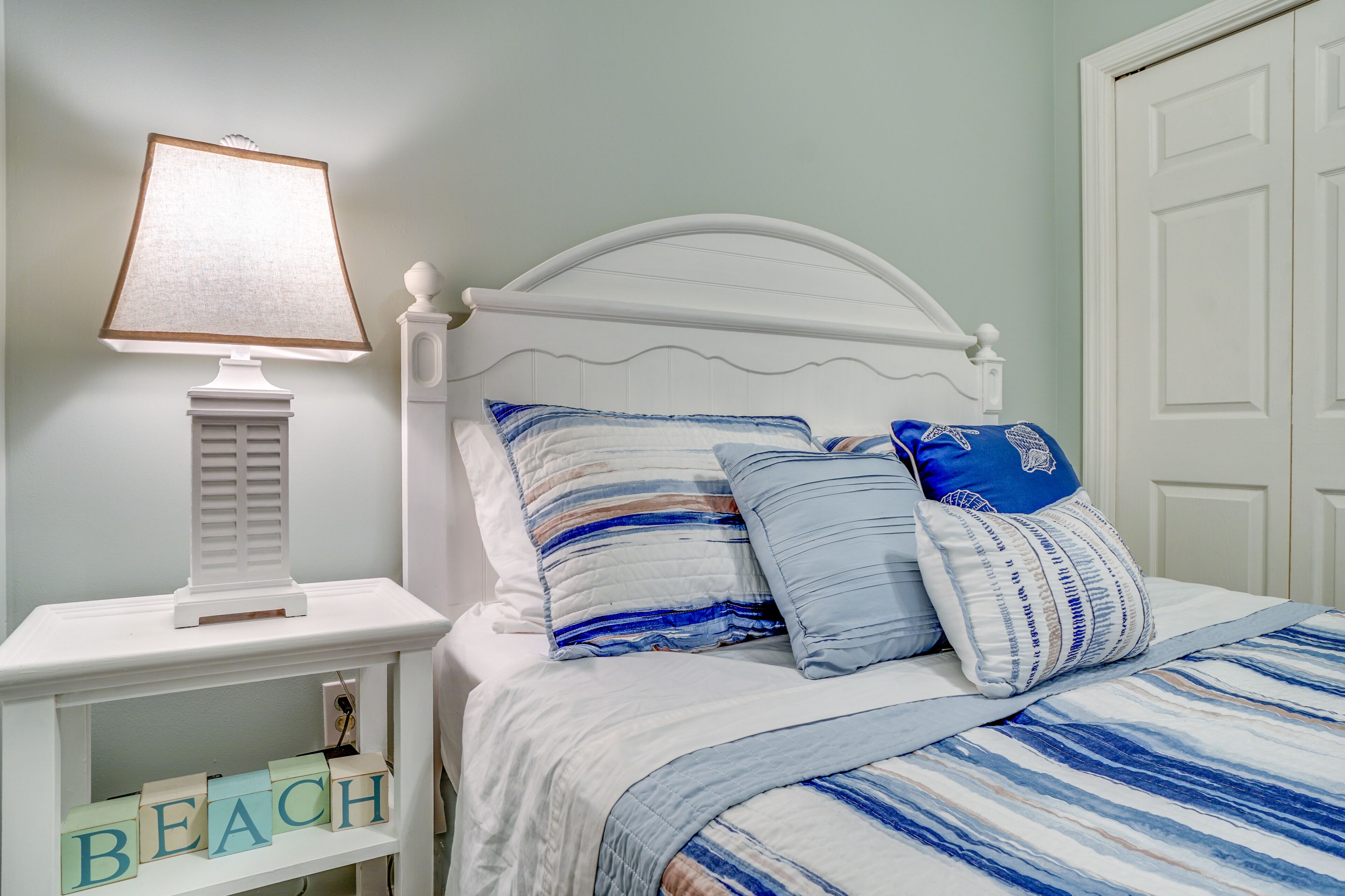 Una cama bien hecha con ropa de cama a rayas, una mesita de noche blanca con una lámpara y un estante con bloques que deletrean "BEACH".